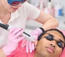 Beauty clinic doctor in nitrile gloves and safety goggles directing laser beam at customer cheek area