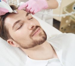 Man resting with his eyes closed while beauty expert moving ultrasonic skin scrubber and removing dirt from pores on forehead