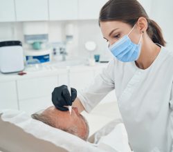 Female beautician wearing medical mask and sterile gloves while cleaning man forehead with cleansing tool